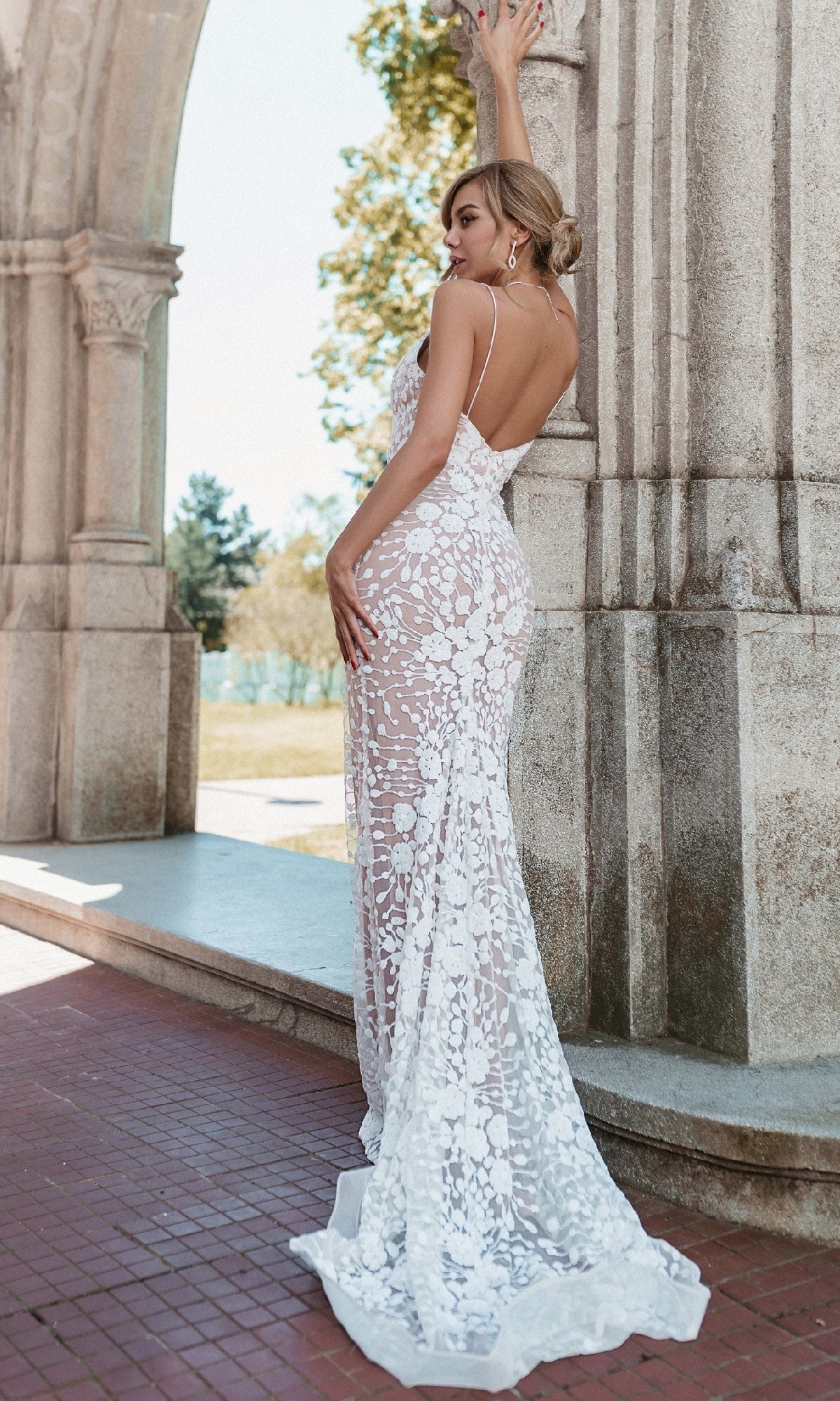 A woman wearing the Long White Lace Formal Dress: Tina Holly BB008 stands under a stone arch, facing away and gently touching the arch with one hand.